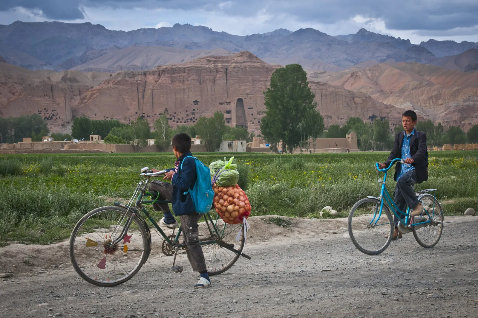 Two local boys cycling up a dirt road in the Bamyan valley, the empty Buddha niche in the cliffs behind